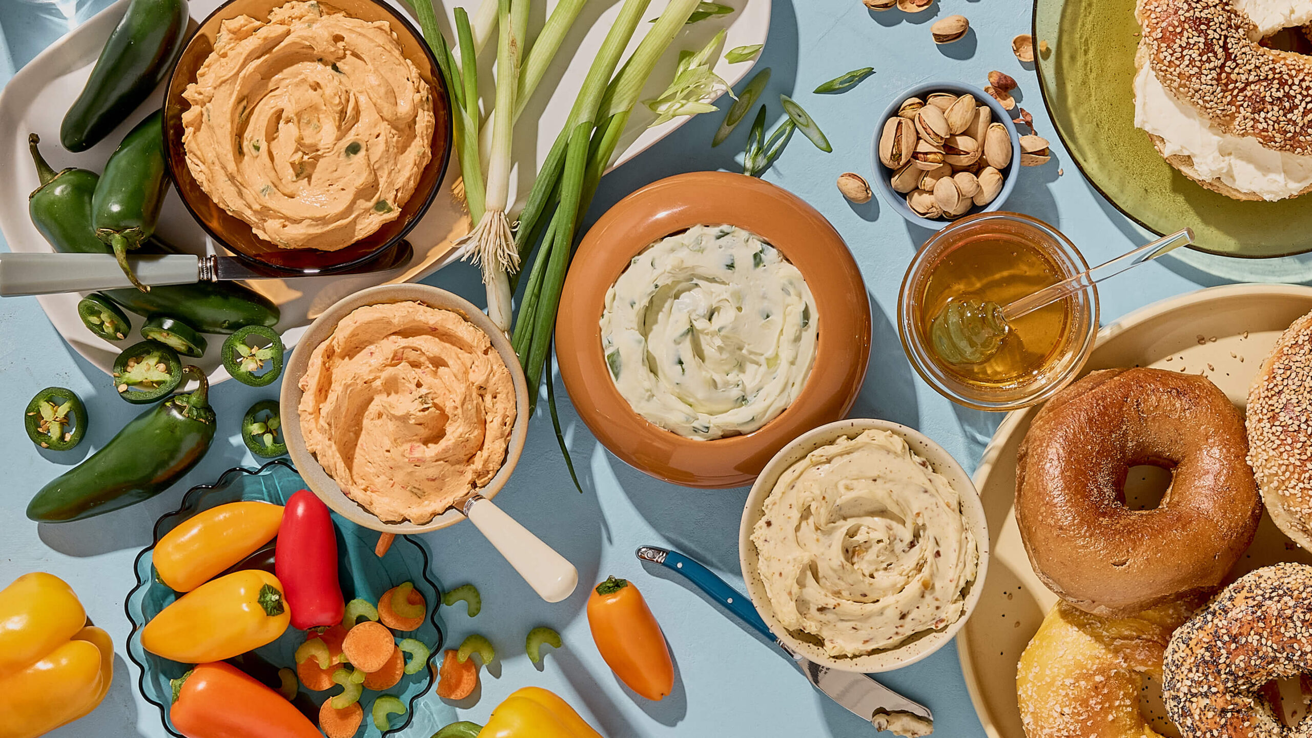 Montreal-style bagels with flavored cream cheeses, fresh peppers, scallions, and honey on a bright blue background.
